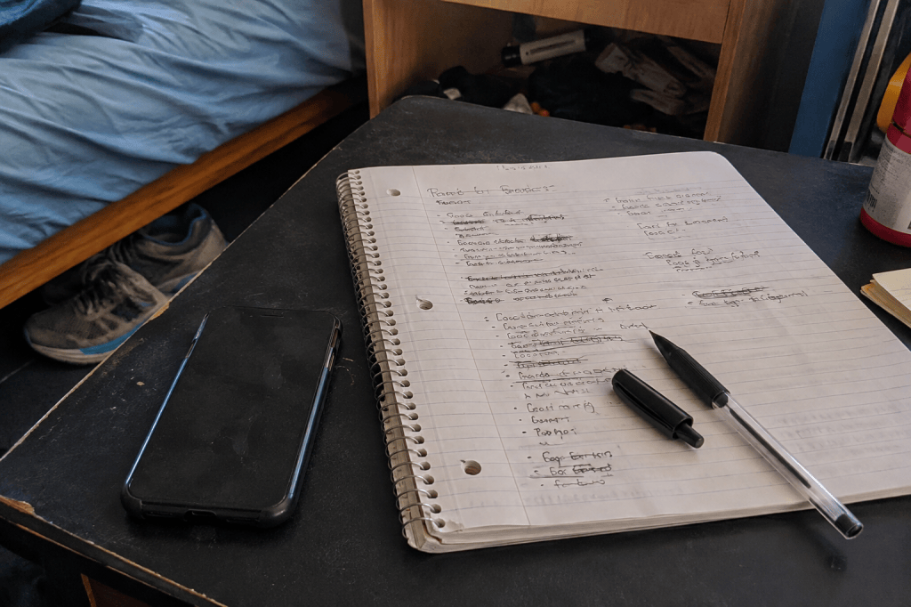 A face-down phone beside a handwritten notebook and pen on a worn dorm-room desk.